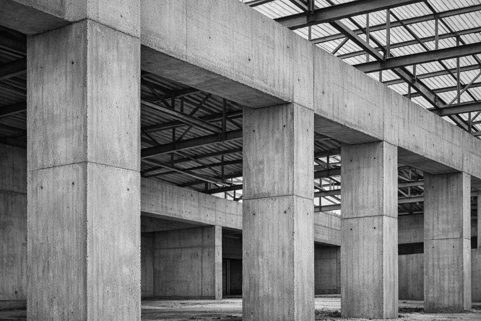 Détail de colonnes en béton et d’une charpente métallique, en noir et blanc.